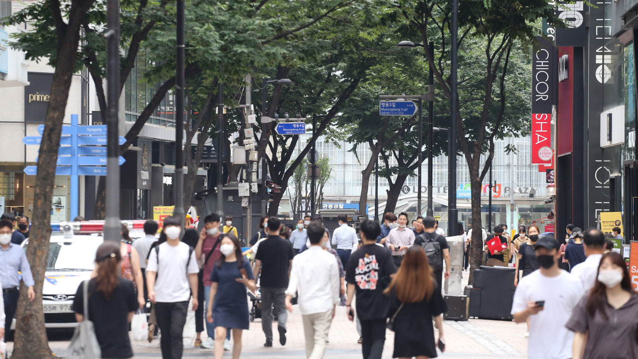 ฝูงชนเดินบนถนนที่เต็มไปด้วยร้านค้าภายใต้ต้นไม้ใหญ่ในเมืองเกาหลีใต้ ขณะสวมหน้ากากอนามัยตามมาตรการป้องกันโควิด-19