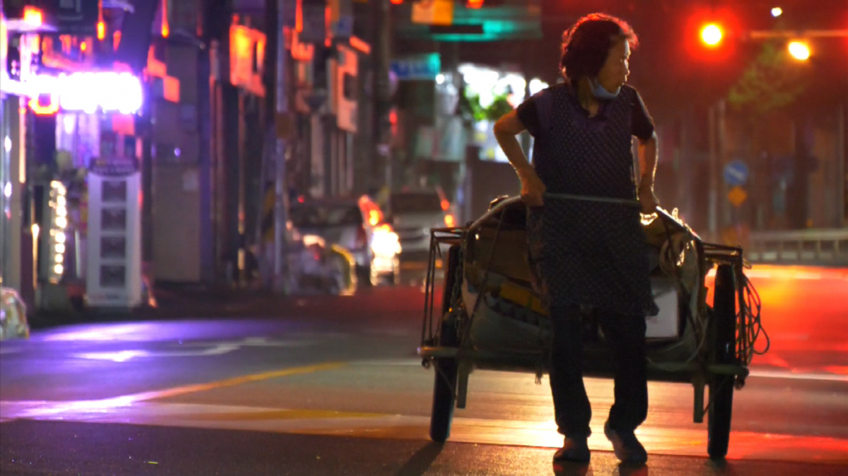 ผู้หญิงคนหนึ่งกำลังดึงรถเข็นผ่านถนนที่มีแสงไฟนีออนติดสว่างในตอนกลางคืน.