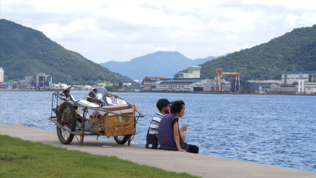 เด็กชายและคุณยายที่ชายฝั่งในชางวอน ประเทศเกาหลีใต้คุยกัน ข้างข้างรถเข็นบรรจุกระดาษและสิ่งรีไซเคิล.