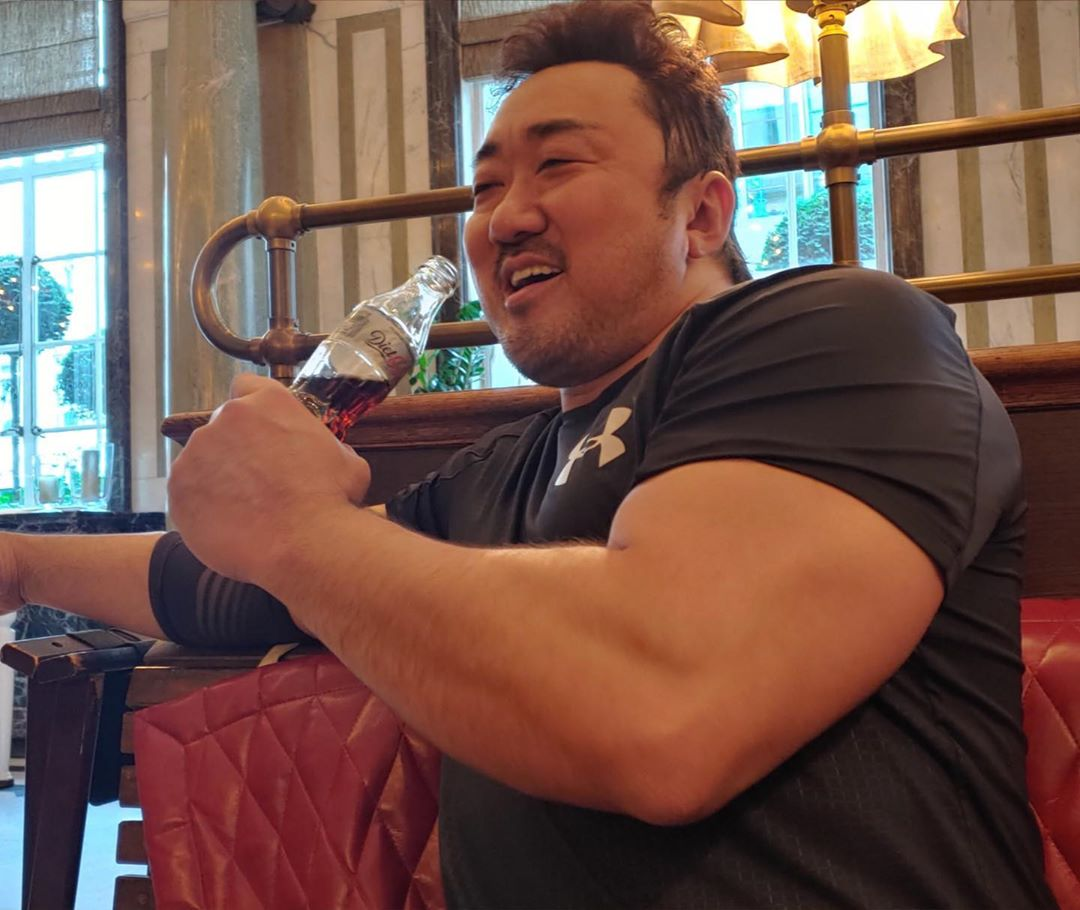 馬東石身穿黑色運動衫，坐在咖啡店裡，肌肉強壯的手臂握著一瓶飲料，展現出他的力量與活力。