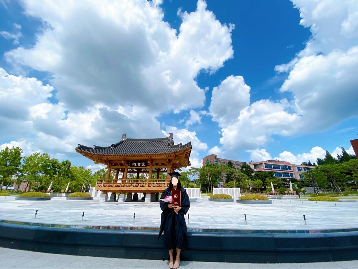 บรรยากาศงานรับปริญญาที่มหาวิทยาลัยในเกาหลีใต้กับท้องฟ้าสดใสและศาลารูปแบบเกาหลีดั้งเดิมในพื้นหลัง ซึ่งนักศึกษาเข้าร่วมพิธีรับปริญญาในบรรยากาศที่ผ่อนคลาย