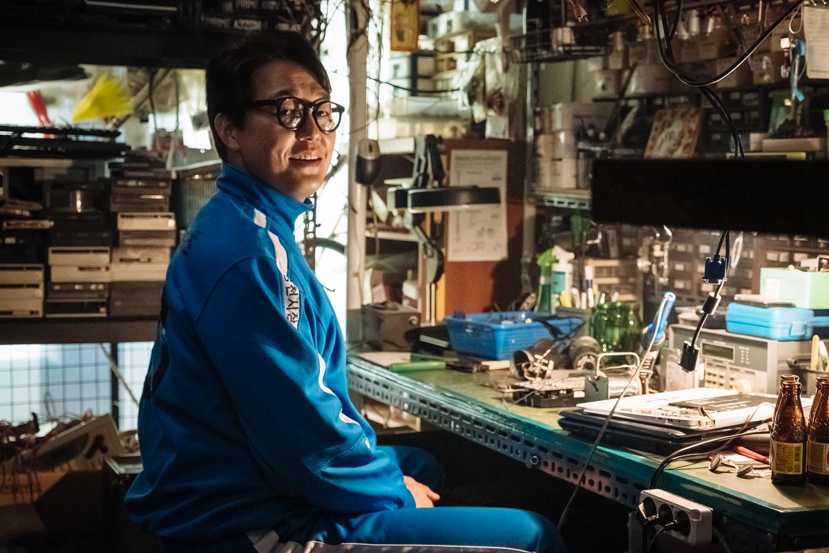 韩国电影《特务搞飞机》中男主角坐在他的工作室，穿着蓝色运动装，光线舒适的环境中摆满了电子设备。