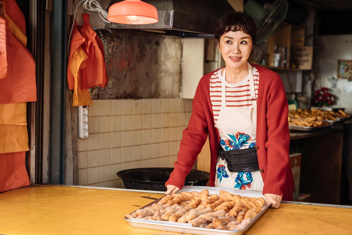 韩国电影《特务搞飞机》中的一幕，女主角站在她的店里，身穿条纹上衣和红色外套，前面是摆满食物的台面。