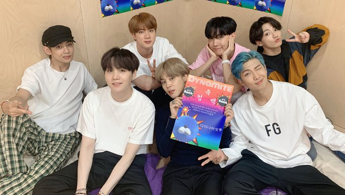 BTS 成員慶祝歌曲 Dynamite 登上 Billboard Hot 100 首位的合照,背景佈置有 Dynamite 宣傳海報,展現喜悦的氛圍