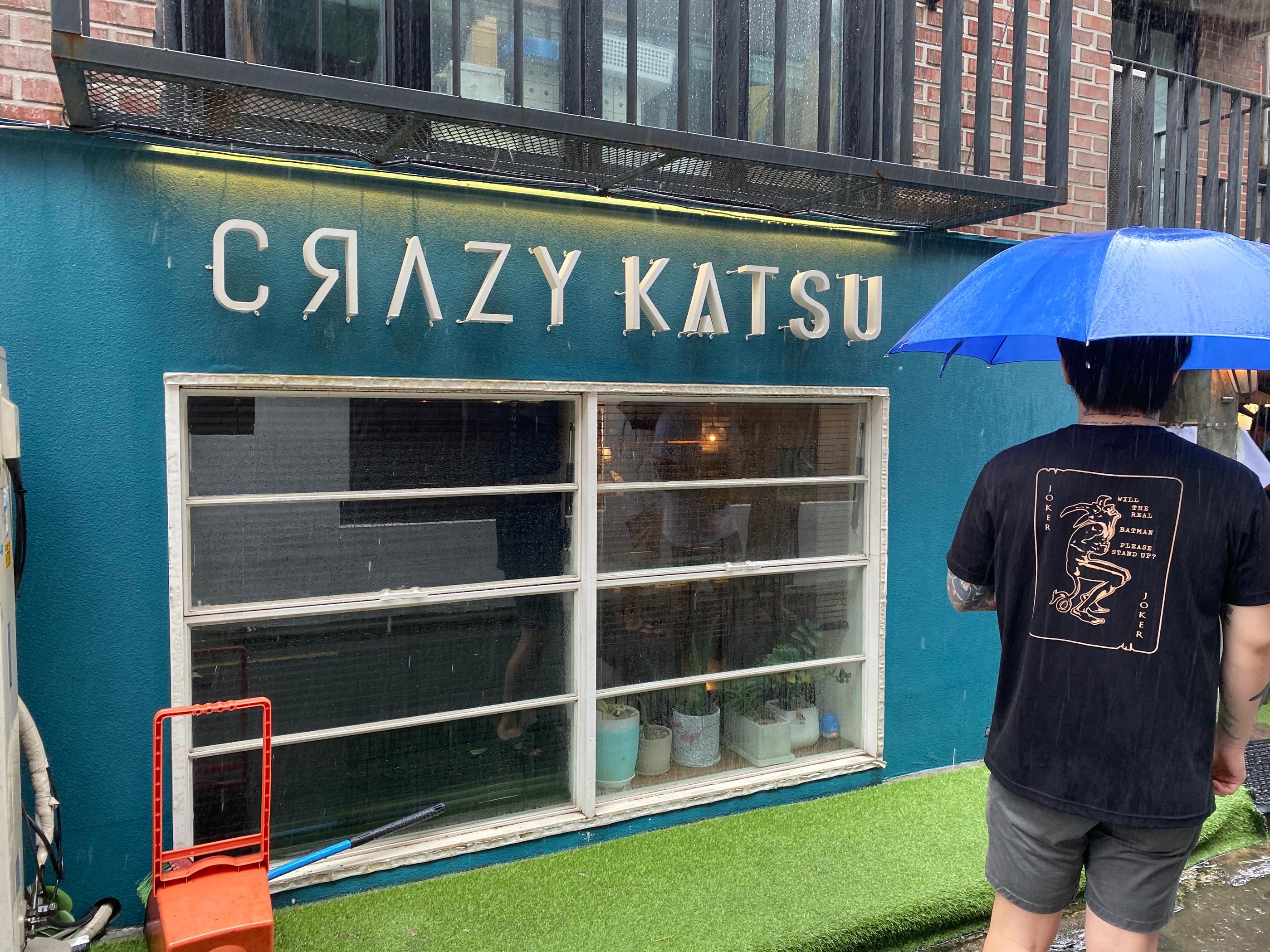 Fachada del restaurante Crazy Katsu en Hapjeong bajo la lluvia, mostrando el cartel iluminado del establecimiento.