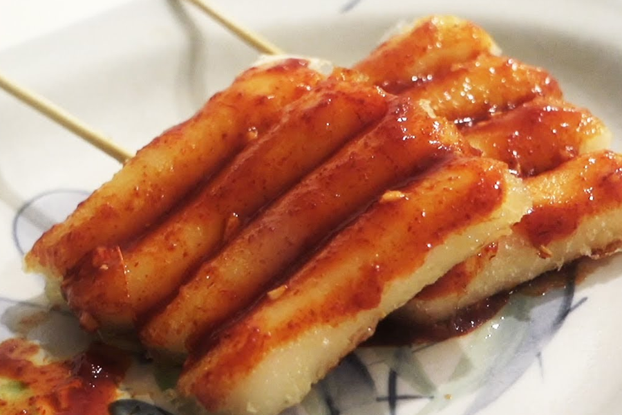 面帶微笑的角色，持有放在竹籤上的韓式辣糕，描述別人的糕好像總是比較的表情夾於上下文之間。