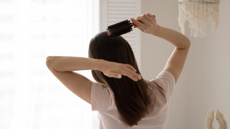 一道門上掛著閃亮的串珠卷簾，對比著日光，房間內有一位穿著淡色T恤的女子，背對著視角，用捲梳輕柔地梳理著頭髮，畫面展現了寧靜的早晨和日常護理的鏡頭。