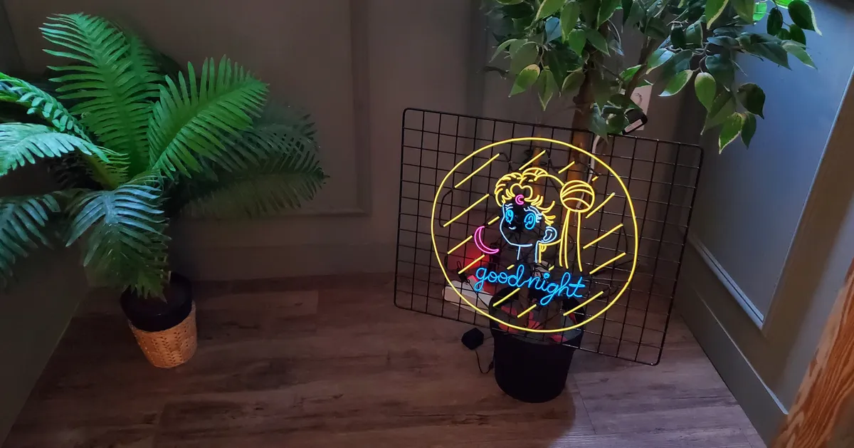 一个光线昏暗的房间里，有一台可爱卡通图案的自制霓虹灯，灯下有青草绿植，营造出温馨的气氛。
