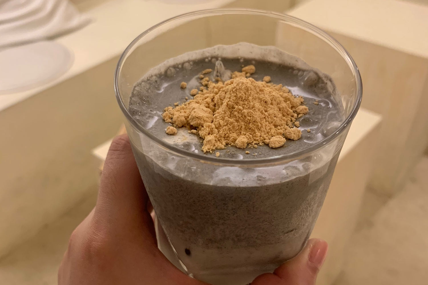 手持一杯黑芝麻拿鐵，飲品上灑有黃豆粉，展示了濃厚香味的咖啡特色。