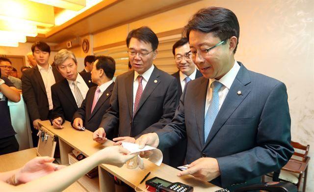 ชายในชุดสูทอยู่ในแถวที่เคาน์เตอร์ชำระเงิน โดยมอบบิลให้กับแคชเชียร์ภายในร้านอาหารแบบเกาหลี ซึ่งแสดงถึงการชำระเงินในร้านอาหารเกาหลียุคปัจจุบัน