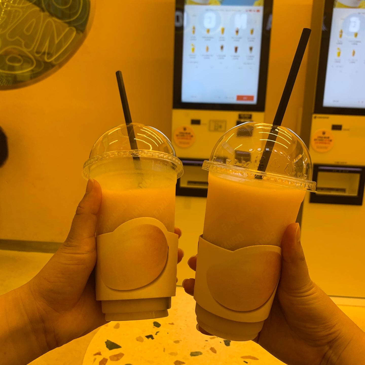 GO MANGO 카페에서 제공하는 망고 음료 두 잔을 손에 들고 있는 모습. 뒷 배경으로 메뉴 스크린이 보임.