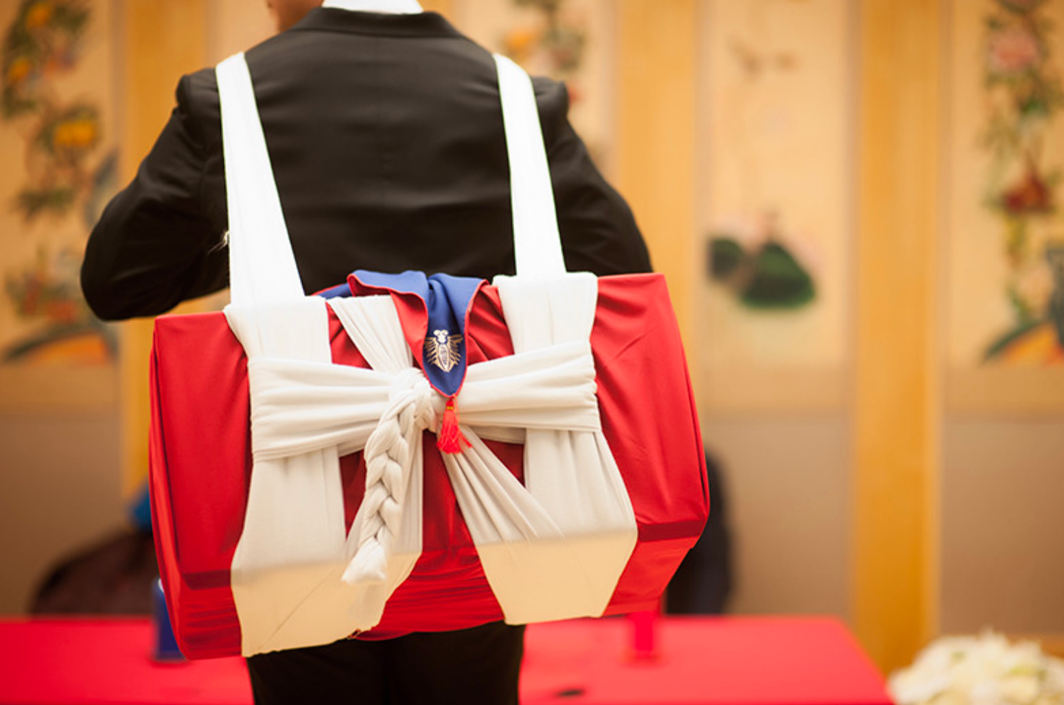 Một người đàn ông mặc vest đang đeo một chiếc hộp lớn, quà tặng hồi môn truyền thống trong đám cưới Hàn Quốc, trông trang trọng và được trang trí bằng vải đỏ và trắng, tượng trưng cho sự tươi đẹp và giàu sang trong cuộc sống vợ chồng.