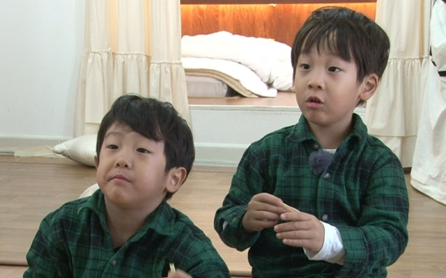 穿著綠色襯衫的雙胞胎坐在地板上，好奇地看著前方，旁邊是柔和的室內環境。