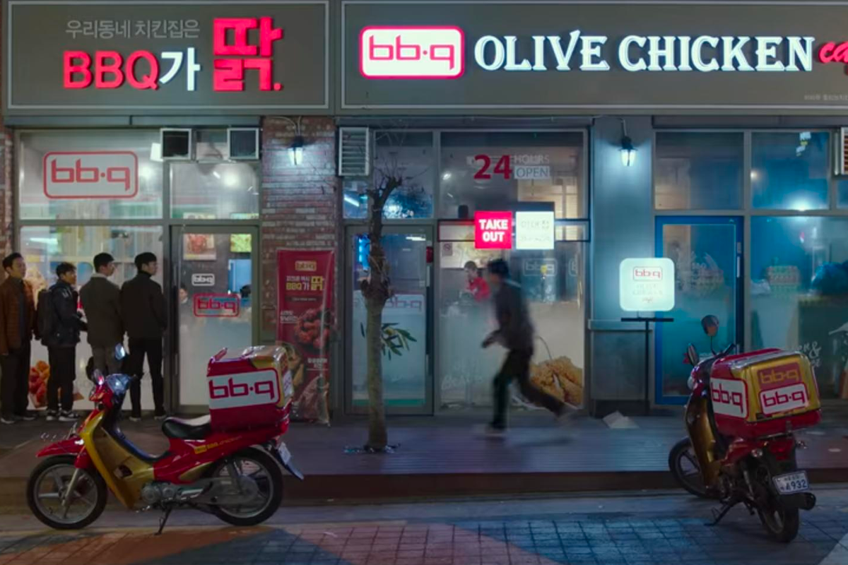 一間韓國知名嘅炸雞品牌BBQ剛烤雞店舖，顧客排隊準備購買炸雞，前面停著兩台外賣電單車。