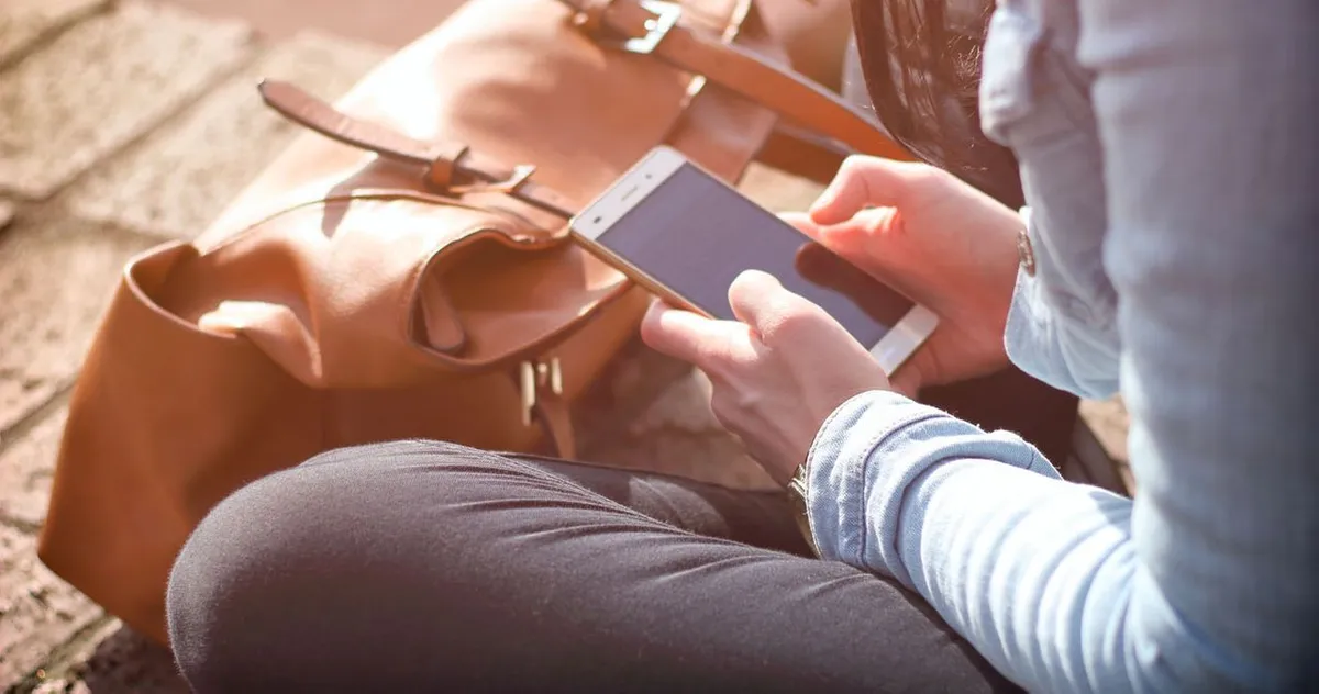 แชทตัวย่อในแชทของคนเกาหลี ภาพผู้หญิงกำลังเล่นโทรศัพท์มือถือ หน้าเธอไม่ชัดเจน อยู่ในที่กลางแจ้ง ข้างกระเป๋าสีน้ำตาล แสดงถึงเทคโนโลยีและการสื่อสารในชีวิตประจำวันของเกาหลี