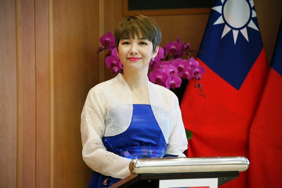 韩国人穿着传统韩服在台湾国庆大会主持活动，背景是两面台湾国旗和紫色花卉装饰，展现多元文化交融。