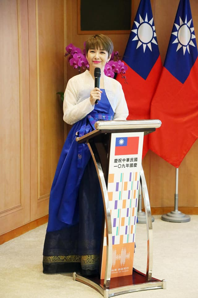 台湾国庆大会上一位穿着韩服的主持人在演讲台前致辞，背景有两面台湾国旗，象征文化多元与国际交流。