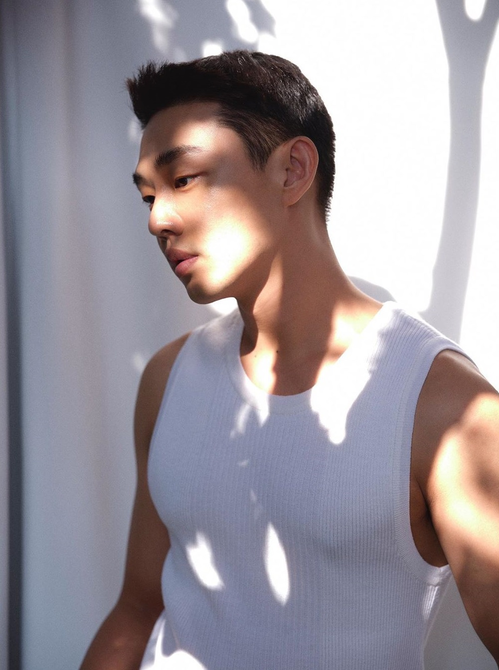 白いタンクトップを着た男性の姿。ヘアスタイルは前髪を上げたアイビーリーグカットのようなスタイルで、サイドは短く刈り上げられている。自然光が身体に影を作り、リラックスした雰囲気を醸し出している。
