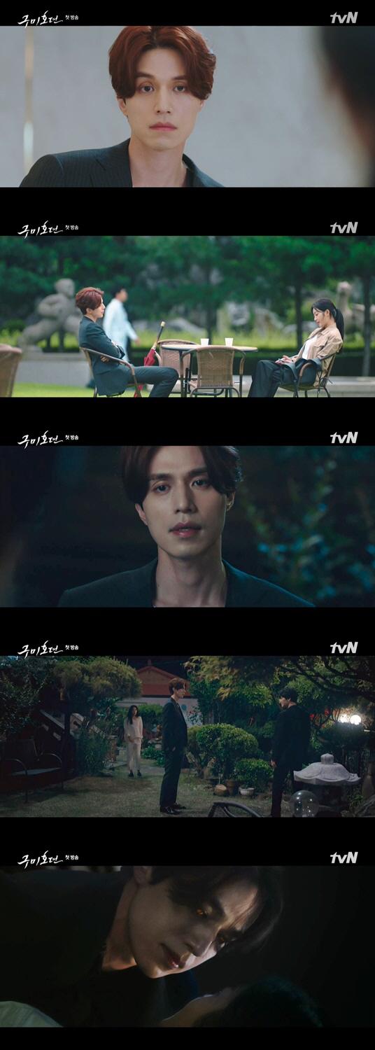 九尾狐伝のシーンで、スーツを着た俳優が片手に赤い傘を持って堂々とした姿で立っている。背景には都市の建物が見える。