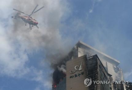 火災の煙が上がる蔚山の高層アパートの上空を飛ぶヘリコプターの写真。避難に関与し、安全性を確保する消防活動の一環として、水を散佈して火を消すために使用される様子が写っている。