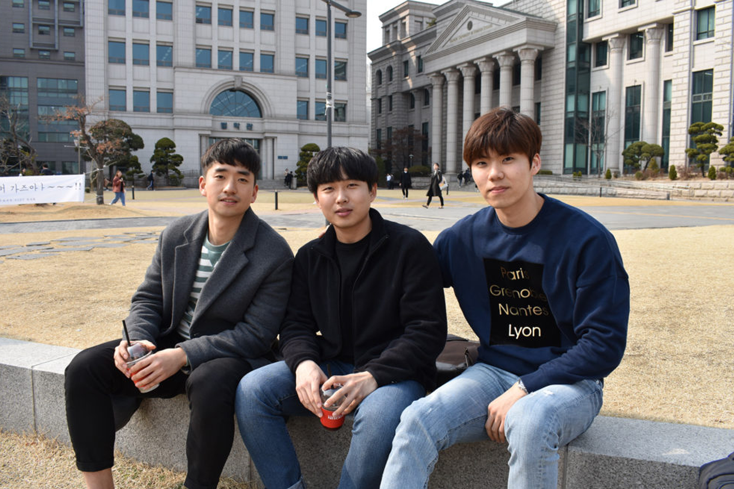三名青年坐在韓國某大學校園長凳上，背景是一座大型建築，地上草坪顏色略有枯黃