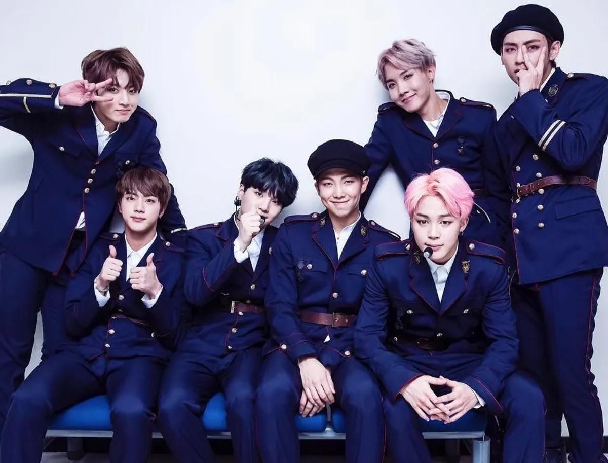 บีทีเอสในชุดสูทสีน้ำเงินเข้มที่ดูเป็นทางการ ขณะนั่งเรียงกันบนโซฟา ฉากหลังเป็นพื้นขาวเรียบ ช่วยเสริมสร้างภาพลักษณ์ที่เข้มแข็งและเป็นทีมเดียวกัน