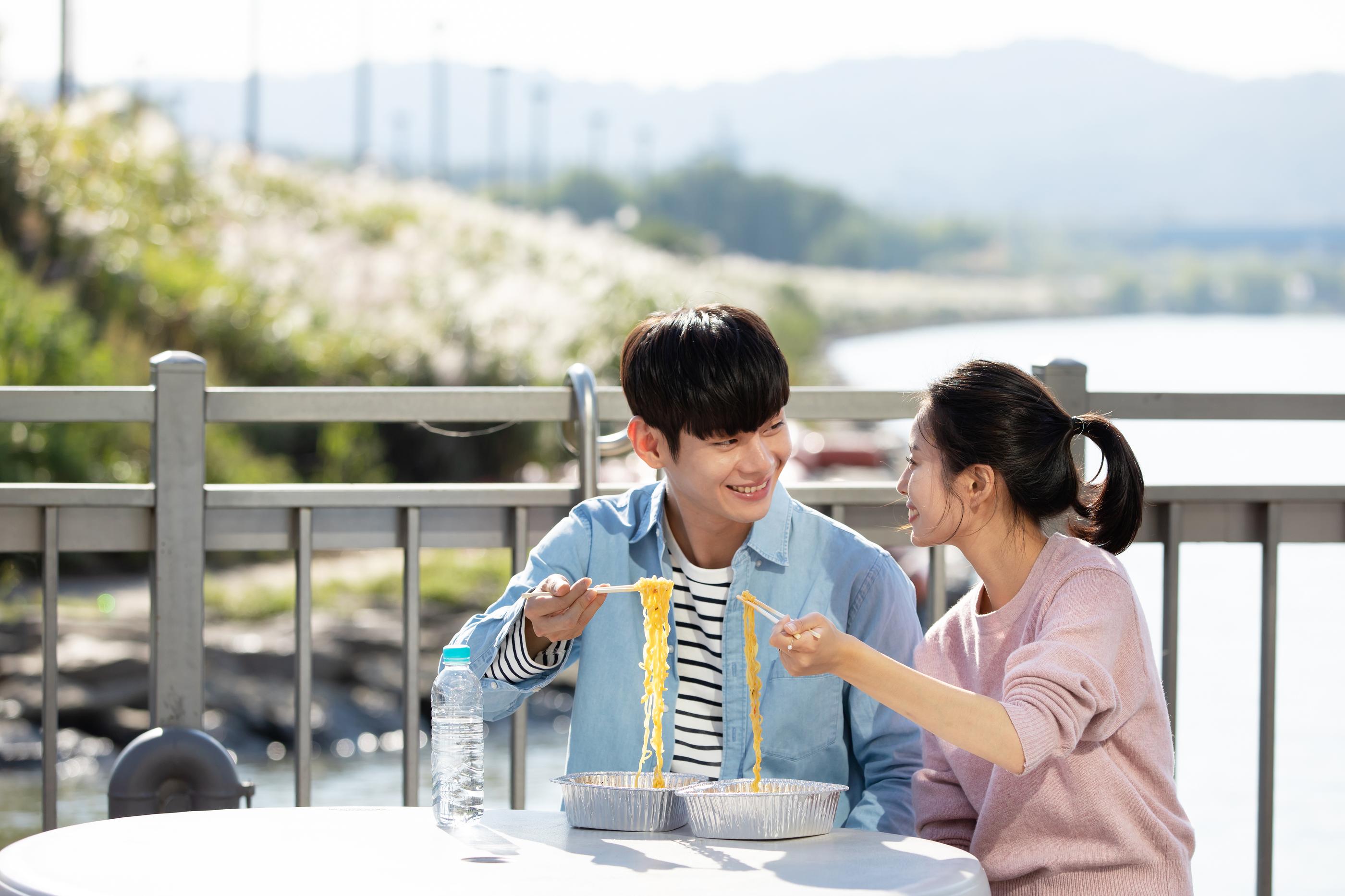 韓国の川沿いでピクニックをするカップル。開放的な景色とともに、テーブルでインスタントラーメンを楽しんでいます。
