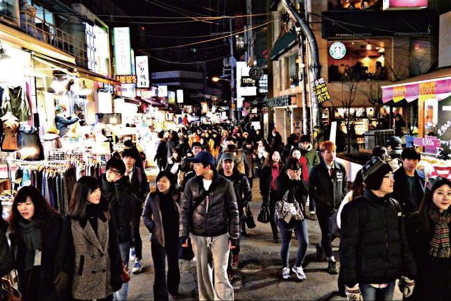韓国の大学生が集まる賑やかな通りでの夜の風景。多くの人々がショッピングや食事を楽しんでいる様子が見え、さまざまな店舗の看板が輝いています。