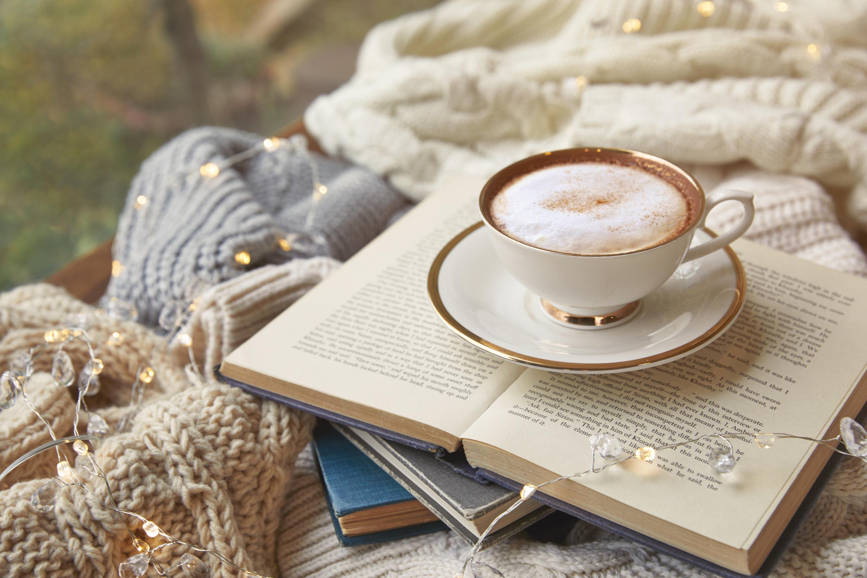 ふわふわのセーターに囲まれた、開いた本とカフェラテが置かれた温かみのあるカフェのテーブル。