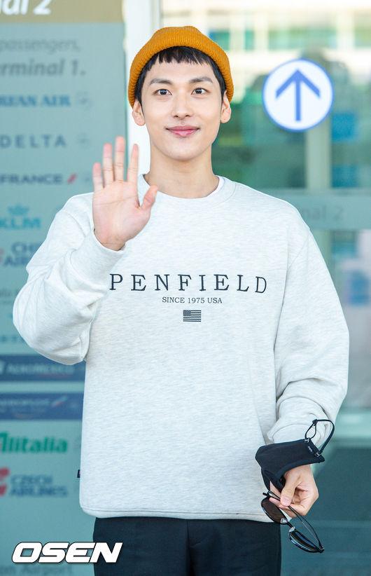 イム・シワンが空港でグレーのスウェットシャツを着て、笑顔で手を振って出発する場面。オレンジ色のビーニーが特徴的。