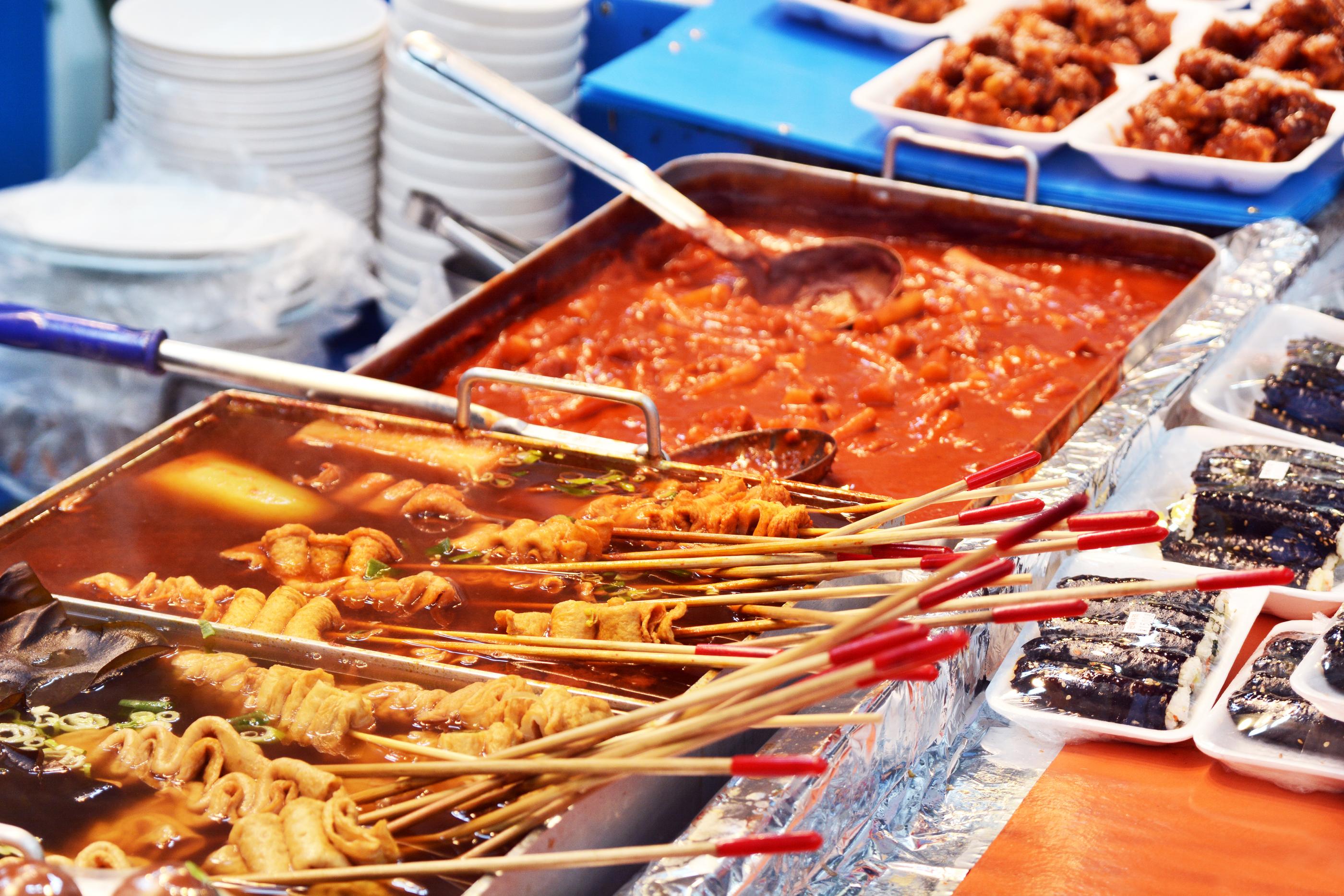 韓国屋台で提供されている料理: トッポッキ、オデン、キムマリの取り合わせ。赤くて甘辛いソースに浸されたトッポッキがメインで、串に刺されたオデンと黒い海苔巻きがサイドに並んでいます。