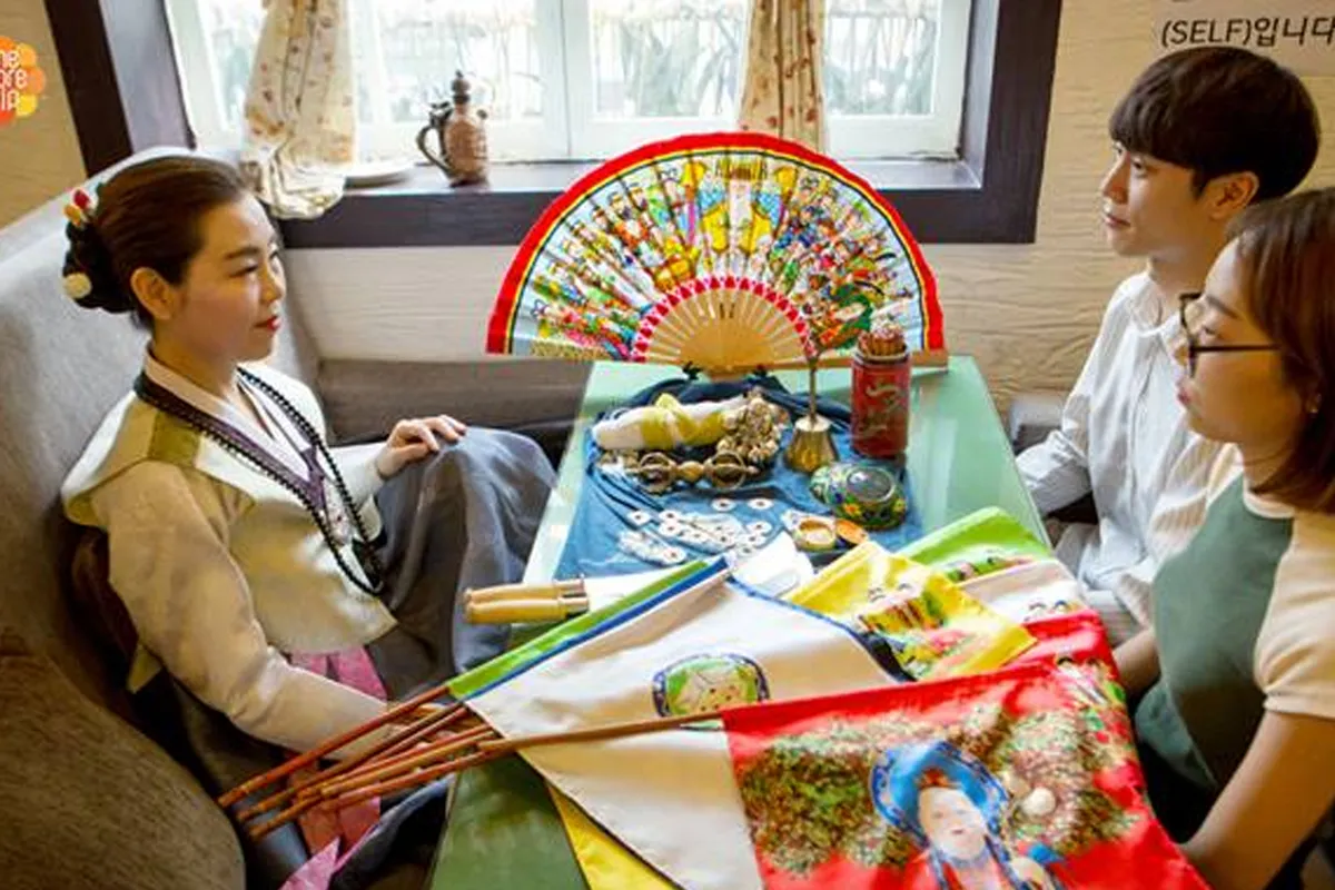 三個坐在桌旁的人物,伴隨著具有傳統韓國文化元素的物品,以占卜和算命程序來觀察改名的背景。