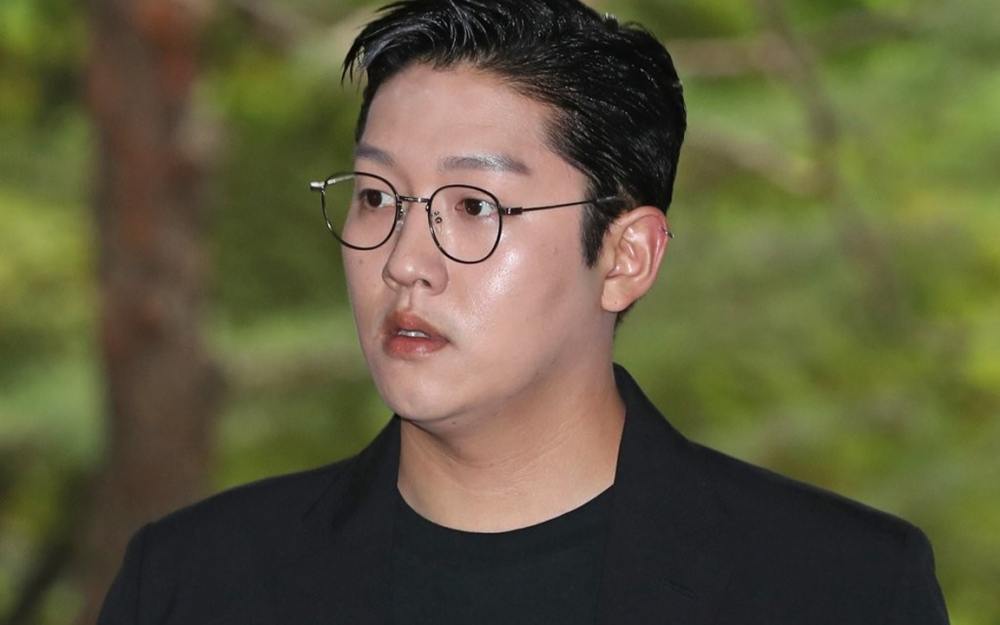 ชายคนหนึ่งสวมสูทสีดำยืนอยู่ท่ามกลางต้นไม้เขียวชอุ่ม