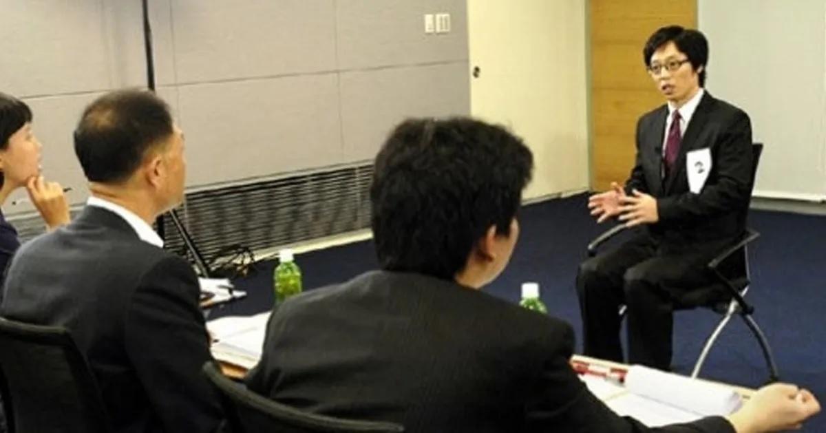 ชายหนุ่มคนหนึ่งกำลังสัมภาษณ์งานกับคณะกรรมการสัมภาษณ์ สามคนในห้องประชุม แสดงถึงขั้นตอนในการหางานที่เป็นไปอย่างแข็งขันในเกาหลีใต้