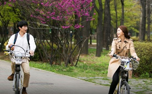 公園裡兩名騎自行車的人在樹下悠閒交談，背景是春天盛開的紫色花卉。