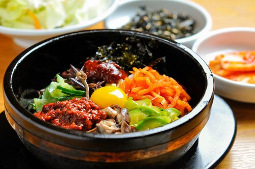 colourful bibimbap served in a black bowl with a golden lining, side dishes, or banchans in the back