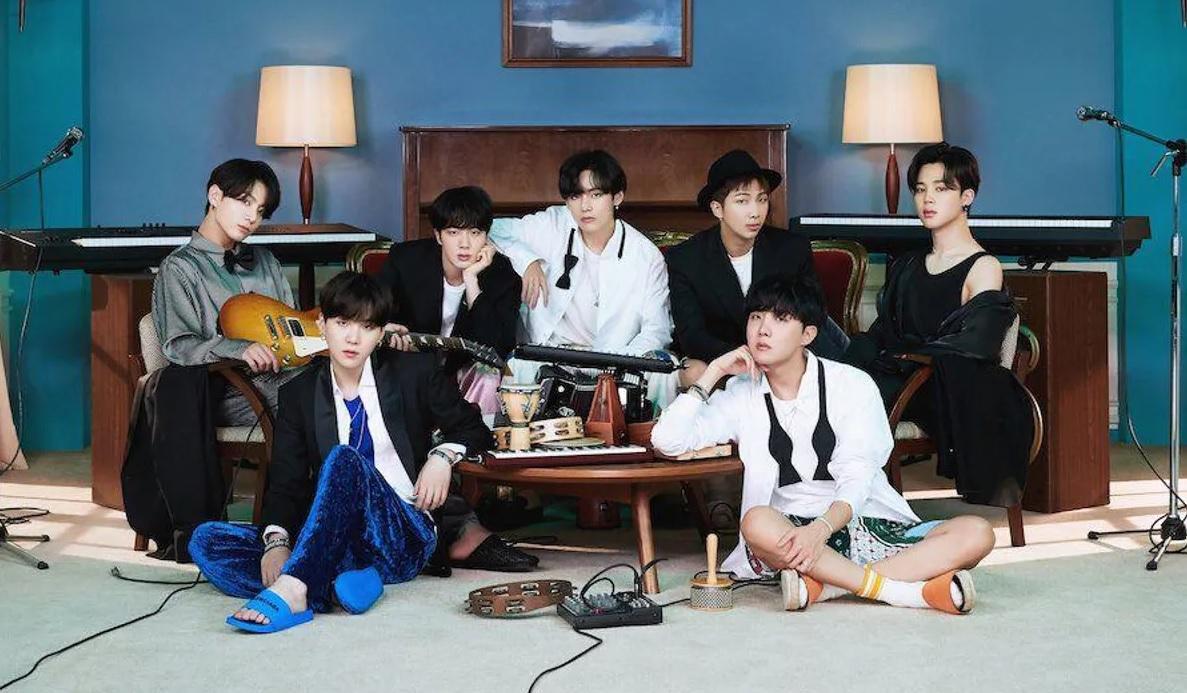 กลุ่มคนเจ็ดคนนั่งรูปในห้องนั่งเล่นตกแต่งด้วยเฟอร์นิเจอร์เก่าและโซฟา หนึ่งในนั้นถือกีตาร์ มีเครื่องดนตรีอื่นๆ วางอยู่รอบๆ พื้นห้องเป็นพรมสีอ่อน ให้บรรยากาศอบอุ่น