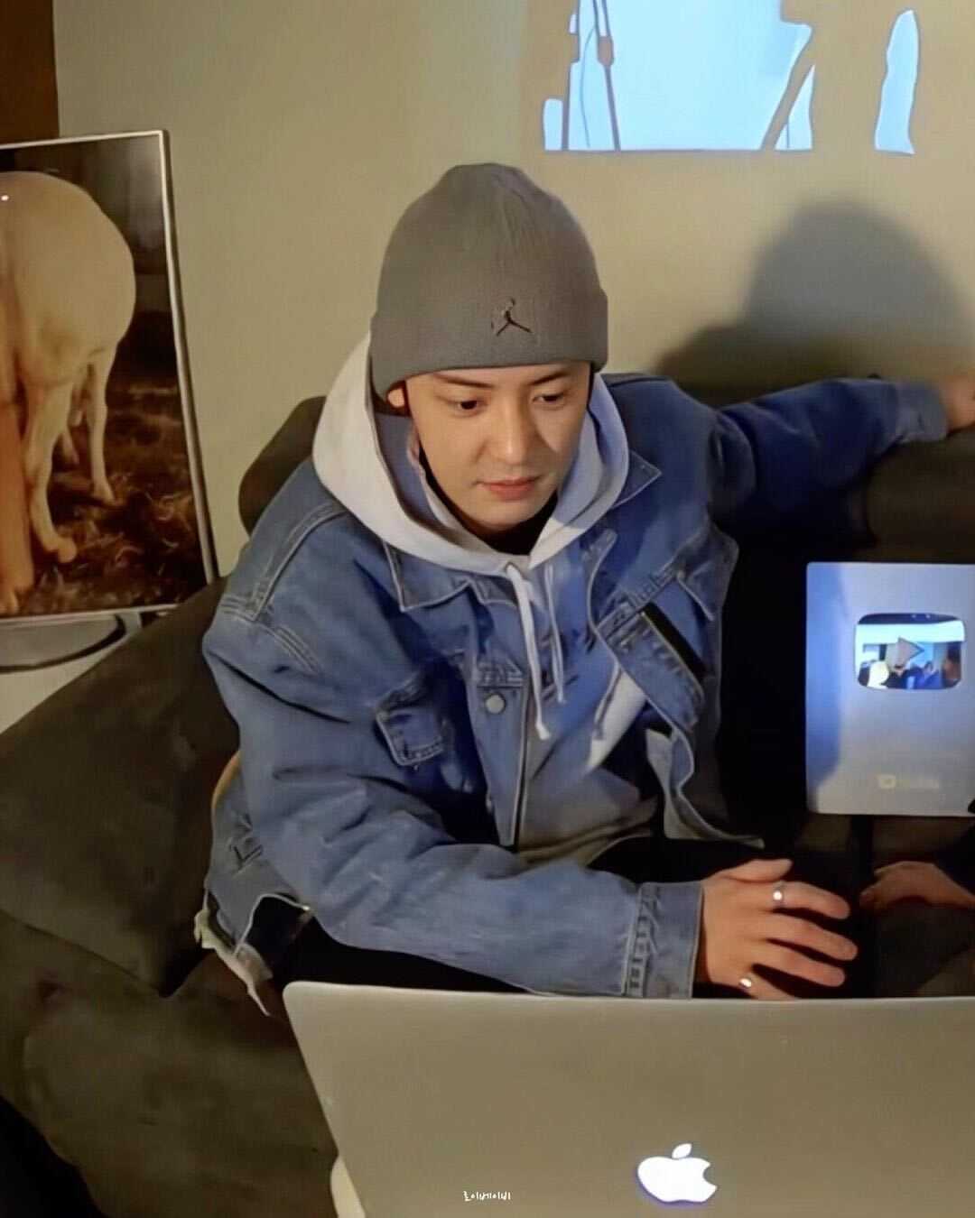 บุคคลสวมหมวกยืนที่โซฟากับแล็ปท็อป Apple ในห้องที่มีโปสเตอร์ตกแต่งผนัง