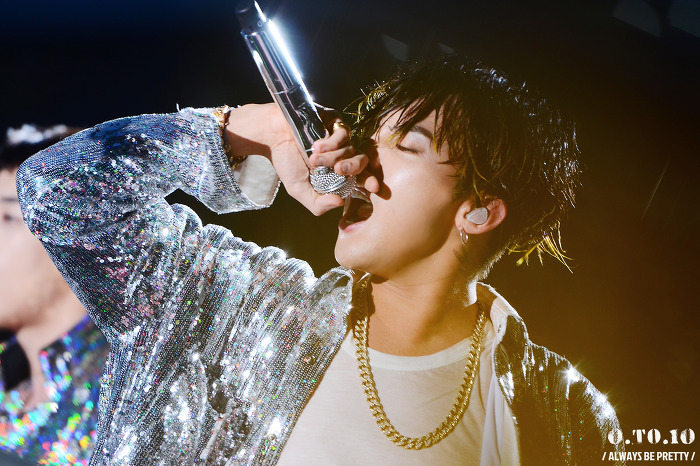 จี-ดรากอน นักร้องหนุ่มจากวง Big Bang กำลังถือไมโครโฟนและใส่เสื้อคลุมแวววาวในการแสดงบนเวที