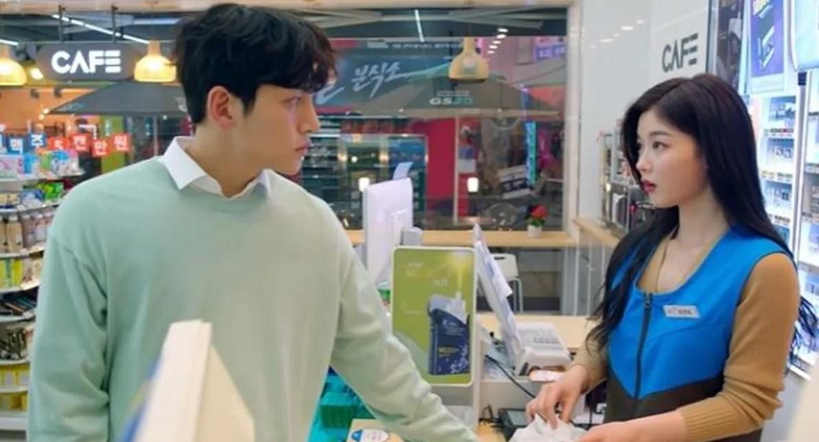 ร้านสะดวกซื้อในเกาหลีใต้, พนักงานร้านสะดวกซื้อหญิงในเสื้อคลุมแขนยาวสีน้ำตาล-ฟ้า กำลังให้บริการลูกค้าชายที่สวมเสื้อกันหนาวสีเขียวอ่อน ซึ่งกำลังรับสินค้า ณ เคาน์เตอร์คิดเงินในร้านสะดวกซื้อที่มีรายการสินค้าหลากหลาย
