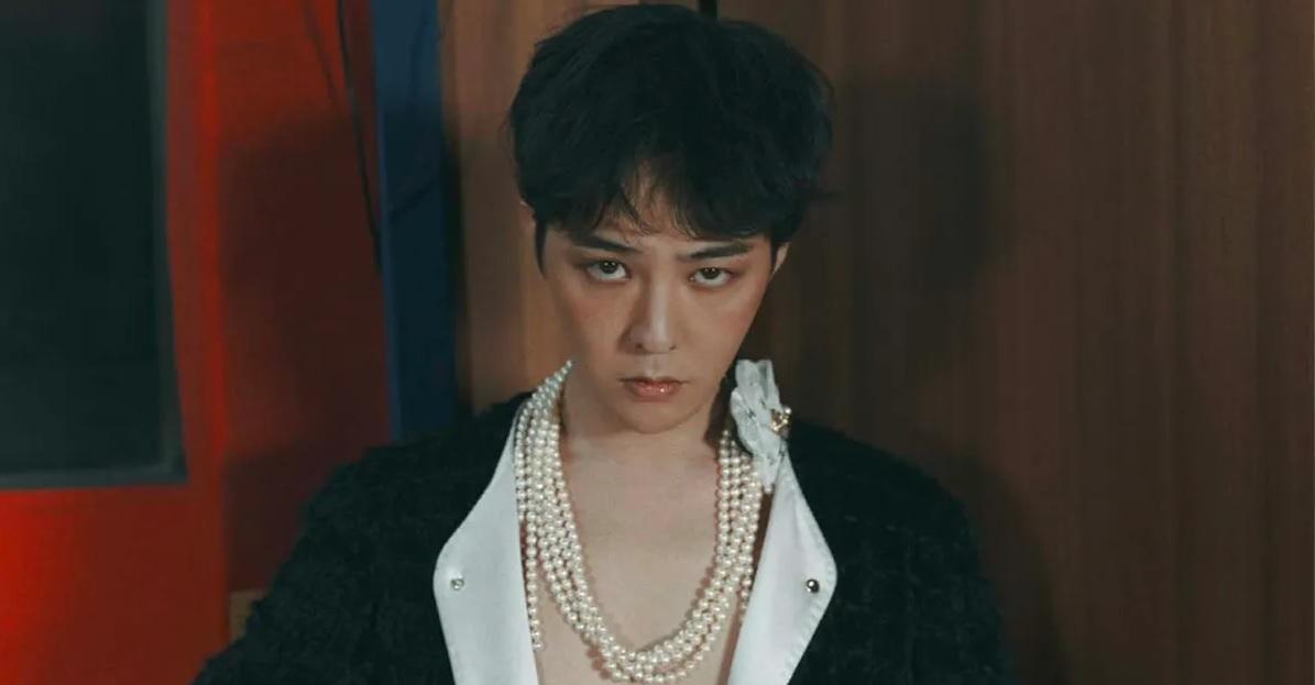 จีดรากอน (G-Dragon) กำลังเตรียมสำหรับการคัมแบ็คใหม่ของเขา พร้อมเสื้อผ้าสีเข้มและสร้อยเพชรด้านหน้า กำลังอยู่ในห้องที่มีบรรยากาศอบอุ่น
