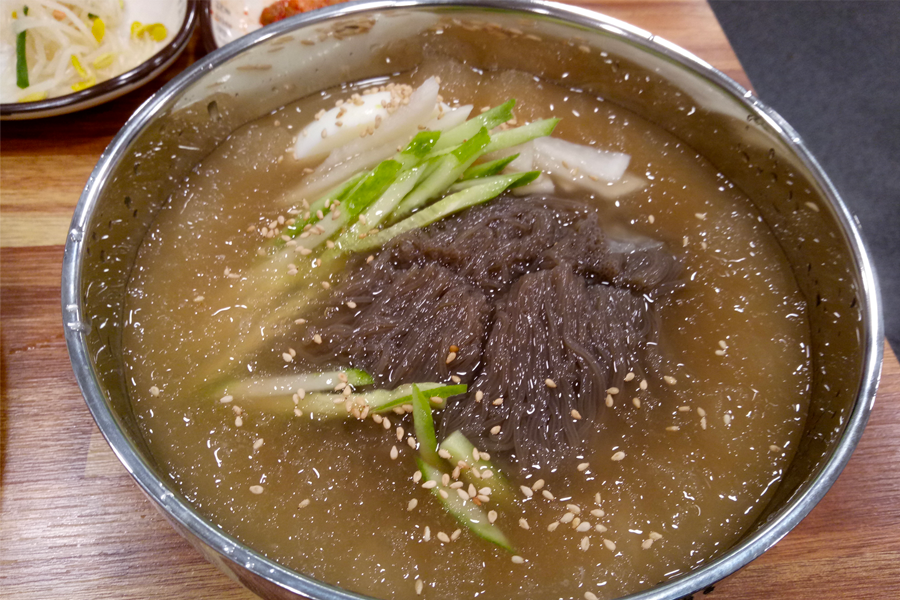 碗裡裝著鹹興冷麵，包含馬鈴薯和地瓜製成的麵條，搭配各種韓國生魚和新鮮香草。