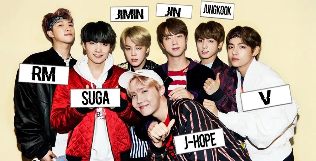 กลุ่มศิลปิน K-pop ยืนเรียงรายกันอยู่พร้อมชื่อในภาษาเกาหลีที่แปะไว้ที่เสื้อของแต่ละคน.