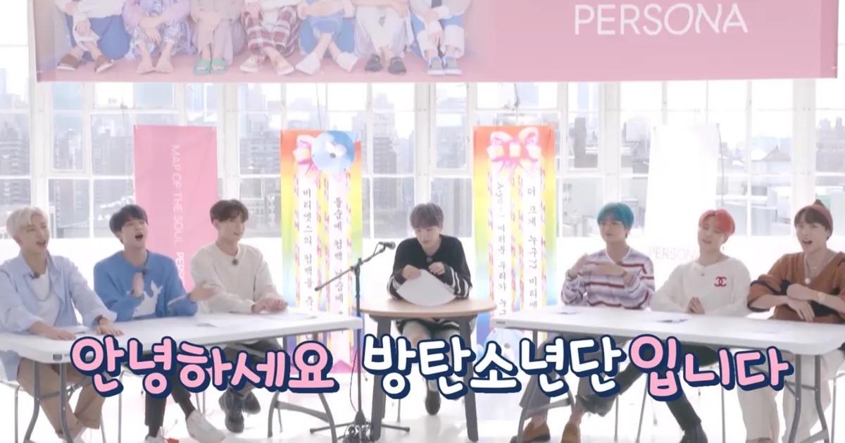 กลุ่มศิลปินนั่งอยู่รอบโต๊ะ โดยมีแบนเนอร์ที่มีข้อความในภาษาเกาหลีและภาพการ์ตูนน่ารักอยู่ด้านหลัง.