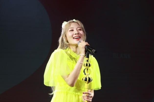 青緑色の髪をしたアン・ジヨンが制服風の衣装を着ている写真。襟に「WORKAHOLIC AN JIYEONG」と書かれた名札がついている。背景は黒で、公式のイベントでの一幕を表している。