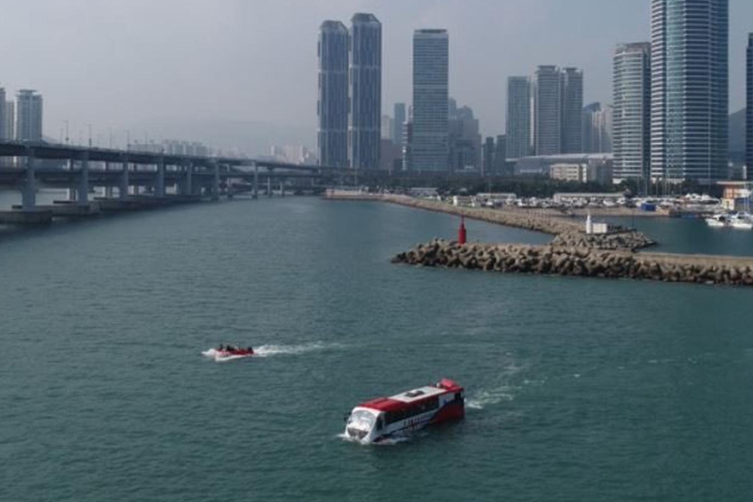 釜山海陸兩用巴士，巴士在水上行駛，遠景是城市天際線和大雄橋。