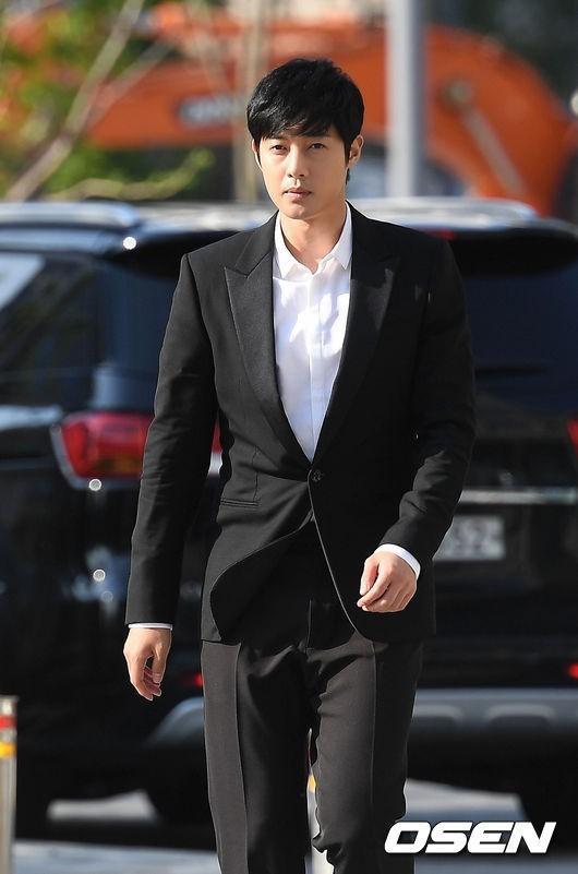 黒いスーツを着て、屋外を歩く俳優。シンプルな白いシャツとスーツスタイル。