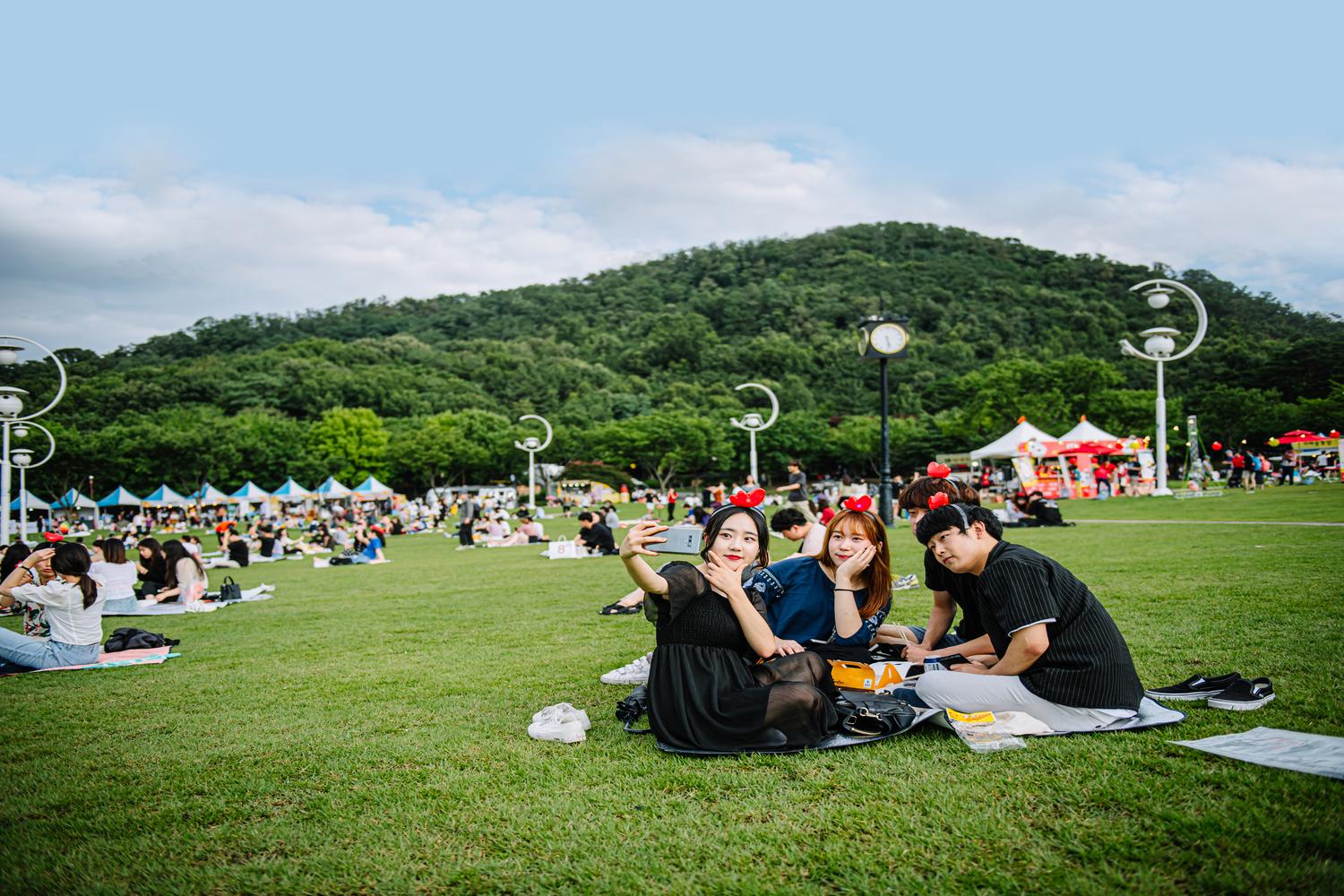 一群人坐在大邱炸鸡啤酒节上的草地上，享受阳光与欢笑，共度美好时光。