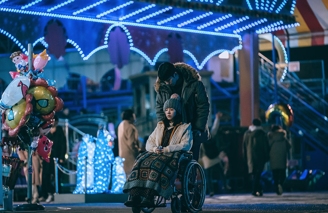 夜間燈火通明的遊樂場，展現出種夢幻的氛圍。男孩推著坐在輪椅上的女孩，兩者間充滿了溫柔的情感。