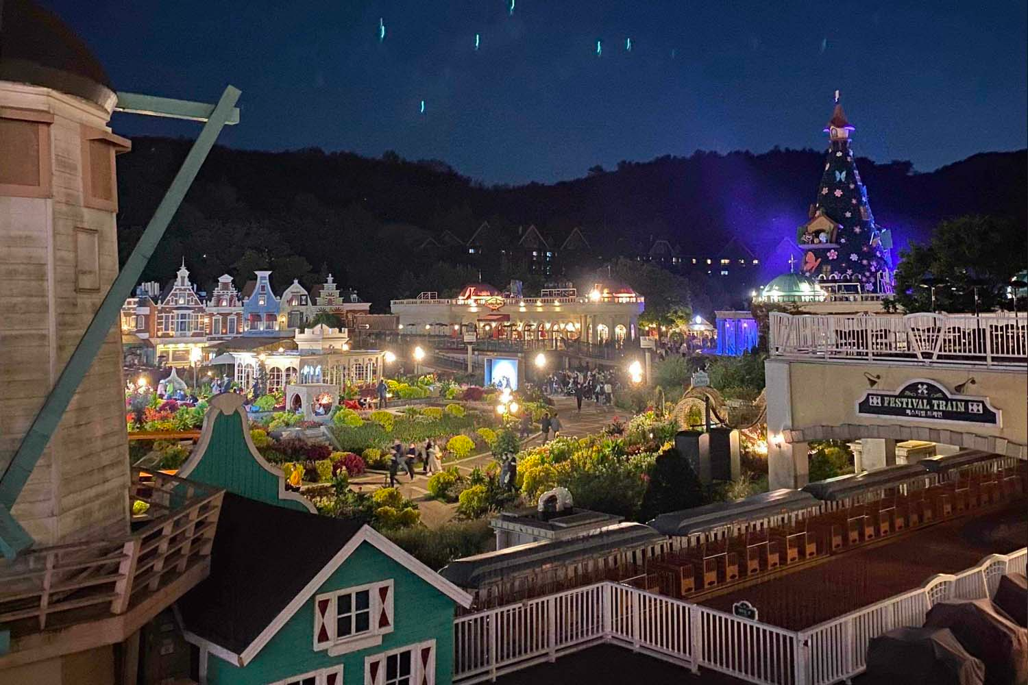 ภาพของสวนสนุกเอเวอร์แลนด์ในเกาหลีใต้ในเวลากลางคืน มองเห็นดอกไม้สีสันสดใสและแสงไฟตกแต่งสวนในบรรยากาศโรแมนติก และมีเทศกาลต้นคริสต์มาสสูงอยู่ตรงกลางภาพ